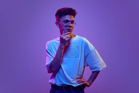 Photo for Young man in casual clothes and glasses standing with thoughtful choosing face against purple background in neon light. Brainstorming. Concept of human emotions, youth, facial expression - Royalty Free Image