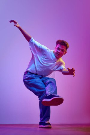 Foto de Joven hombre alegre en ropa casual, jeans y camiseta blanca bailando hip hop sobre fondo púrpura en luz de neón. Hobby, estilo de vida activo. Concepto de emociones humanas, cultura juvenil, moda - Imagen libre de derechos