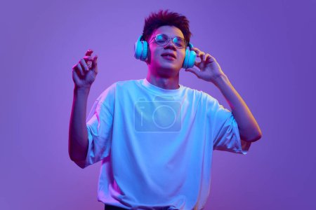 Téléchargez les photos : Jeune homme en t-shirt blanc et lunettes écoutant de la musique dans des écouteurs sur fond violet sous la lumière du néon. Le plaisir. Concept d'émotions humaines, jeunesse, musique, gadgets modernes, activité de loisirs - en image libre de droit