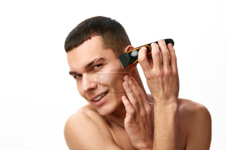 Téléchargez les photos : Beau jeune homme torse nu utilisant un rasoir électrique pour raser le visage sur fond de studio blanc. Bonjour les soins de la peau. Concept de beauté masculine, soins de la peau, spa, cosmétologie, santé masculine - en image libre de droit