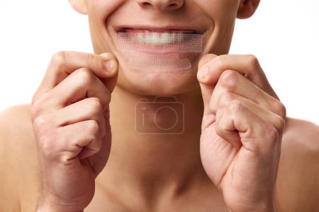 Photo for Cropped image of male face against white studio background. Man whitening teeth with whitening strips. Concept of male beauty, dental care, spa, cosmetology, mens health - Royalty Free Image