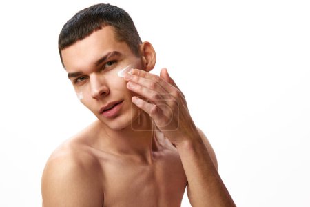 Photo for Handsome young man with short hair applying face moisturizing cream against white studio background. Organic products. Concept of male beauty, skin care, spa, cosmetology, mens health - Royalty Free Image