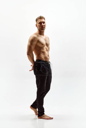 Foto de Imagen completa de un joven guapo con cuerpo musculoso sin camisa en pantalones aislados sobre fondo blanco del estudio. Concepto de belleza masculina, cuidado corporal, fitness, deporte, salud - Imagen libre de derechos