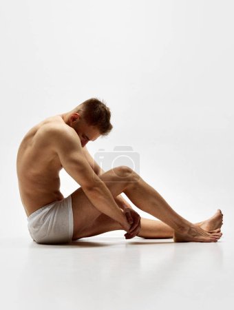Photo for Shirtless young man with fit, sportive, relief body shape sitting on boxers, underwear isolated over white studio background. Concept of male beauty, body care, fitness, sport, health - Royalty Free Image
