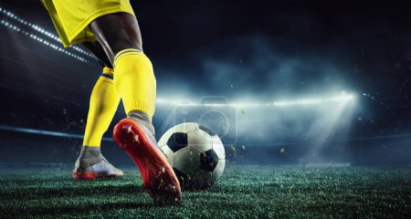 Foto de Imagen recortada de las piernas del hombre africano, jugador de fútbol en uniforme amarillo en la arena 3d jugando, golpeando la pelota. Fiesta nocturna al aire libre. Concepto de deporte, juego, competición, campeonato. Renderizado 3D - Imagen libre de derechos