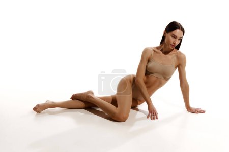 Photo for Beautiful young woman with relief slim fit body sitting on floor, posing in beige cotton underwear over white studio background. Concept of female beauty, body and skin care, nutrition, sport, health - Royalty Free Image