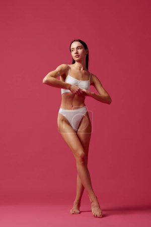 Photo for Body-positivity. Full-length image of young beautiful woman with fit, slim, healthy body in white cotton underwear against pink studio background. Concept of beauty, body and skin care, cosmetology - Royalty Free Image