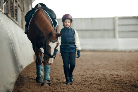 Foto de Niña, niño con uniforme especial y casco caminando con caballo durante el curso educativo de equitación. Comportamiento y cuidado animal. Deporte, infancia, escuela, estilo de vida activo, concepto de hobby - Imagen libre de derechos