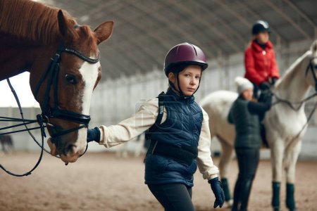 Foto de Niña, niño en casco sosteniendo el caballo y caminando en la arena especial. Curso de equitación, entrenamiento para niños. Concepto de deporte, infancia, escuela, curso, estilo de vida activo, hobby - Imagen libre de derechos
