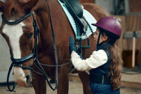 Foto de Niña, niña con casco cuidando a caballo, mostrando su amor. Entrenamiento infantil a caballo en arena especial, pabellón. Concepto de deporte, infancia, escuela, curso, estilo de vida activo, hobby - Imagen libre de derechos