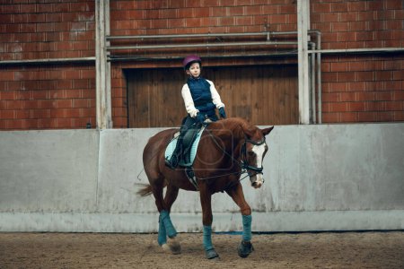 Photo for Little girl, child in uniform and helmet sitting on horse, raining, learning horseback riding activity on special arena, pavilion. Concept of sport, childhood, school, course, active lifestyle, hobby - Royalty Free Image