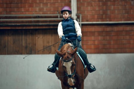 Foto de Niña, niño en uniforme y casco sentado a caballo, lloviendo, aprendiendo a montar a caballo actividad en arena especial, pabellón. Concepto de deporte, infancia, escuela, curso, estilo de vida activo, hobby - Imagen libre de derechos