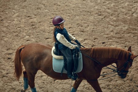 Photo for Concentrated little girl in inform and special clothes siting on horse, training horseback riding on special manege. Concept of sport, childhood, school, course, active lifestyle, hobby - Royalty Free Image