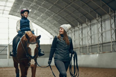 Foto de Mujer joven, maestra, instructora enseñando a una niña a montar a caballo. Niña sentada en remo, practicando. Educación. Concepto de deporte, infancia, escuela, curso, estilo de vida activo y hobby - Imagen libre de derechos