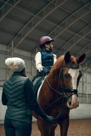 Foto de Instructora enseñando a una niña, a un niño montando a caballo. Niño practicando trail riding, dressage. Concepto de deporte, infancia, escuela, curso, estilo de vida activo, hobby - Imagen libre de derechos