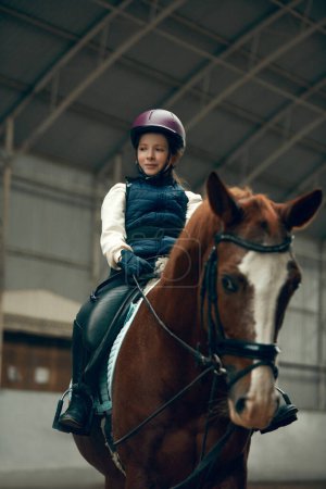 Foto de Retrato de niña, niño en casco sentado en caballo marrón, entrenamiento, práctica de montar a caballo. Concepto de deporte, infancia, escuela, curso, estilo de vida activo, hobby - Imagen libre de derechos