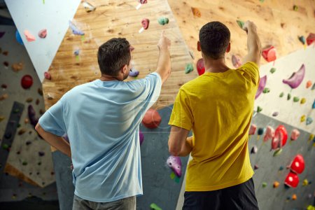 Photo for Man standing with instructor, looking and pointing on climbing wall, discussing techniques of climbing, bouldering activity. Concept of sport climbing, hobby, active lifestyle, school, training course - Royalty Free Image