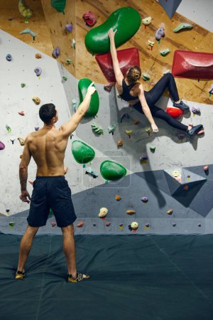 Photo for Muscular shirtless young man, bouldering training teaching young girl to climb wall. Sportive meeting. Concept of sport, bouldering, sport climbing, hobby, active lifestyle, school, training course - Royalty Free Image