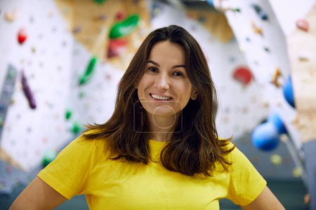Foto de Retrato de una joven mujer sonriente en camiseta amarilla de pie con un muro de escalada detrás. Concepto de deporte, bouldering, escalada deportiva, hobby, estilo de vida activo, escuela, curso de entrenamiento - Imagen libre de derechos