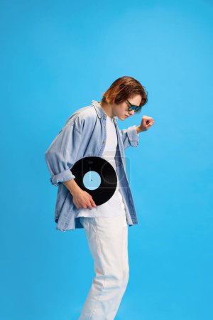 Photo for Side view portrait of happy, overjoyed man, student in modern sunglasses dancing with vinyl record against blue studio background. Concept of retro music and dance, self-expression, party, fashion. - Royalty Free Image