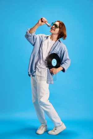 Photo for Young man wearing fashion casual outfit and sunglasses singing retro microphone and holding vintage vinyl record disk against blue studio background. Concept of music and dance, self-expression, party - Royalty Free Image