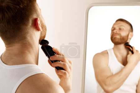 Téléchargez les photos : Homme sérieux rasez la barbe pour l'hygiène, le nettoyage et l'épilation avec un rasoir électrique devant le miroir à la maison. Concept de beauté naturelle, hygiène, soins personnels, soins du visage masculin. Publicité - en image libre de droit