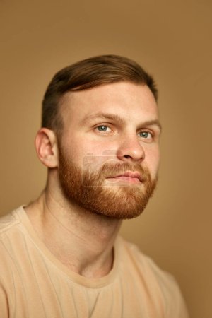 Téléchargez les photos : Portrait de jeune homme roux beau avec barbe en T-shirt beige regardant loin sur fond de studio sablonneux. Concept de soins de beauté, d'autosoin et d'hygiène, cosmétique, spa. Publicité - en image libre de droit