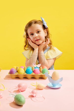 Photo for Portrait of beautiful little girl, child with bow hair accessories sitting at table with Easter eggs against yellow background. Concept of Easter holiday, celebration, traditions, childhood, happiness - Royalty Free Image
