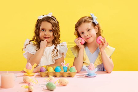 Téléchargez les photos : Petites filles mignonnes, enfants en robes assises à table avec des œufs de Pâques peints sur fond jaune. Suivant les traditions. Concept de vacances de Pâques, célébration, enfance, bonheur - en image libre de droit