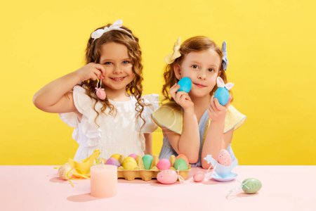 Photo for Beautiful little girls, sisters in dresses holding painted and decorated Easter eggs against yellow background. Concept of Easter holiday, celebration, traditions, childhood, happiness - Royalty Free Image