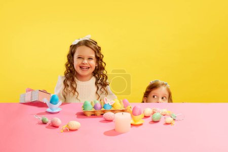 Téléchargez les photos : Joyeux petite fille tenant présent, enfant regardant la table avec des œufs peints colorés peints sur fond jaune. Concept de vacances de Pâques, célébration, traditions, enfance, bonheur - en image libre de droit