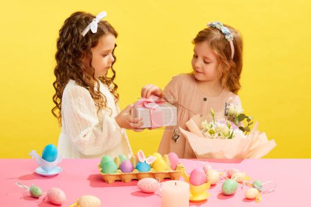 Téléchargez les photos : Petites filles en robe de fête tenant boîte cadeau, fleurs, debout à table avec des œufs peints sur fond jaune. Concept de vacances de Pâques, célébration, traditions, enfance, bonheur - en image libre de droit