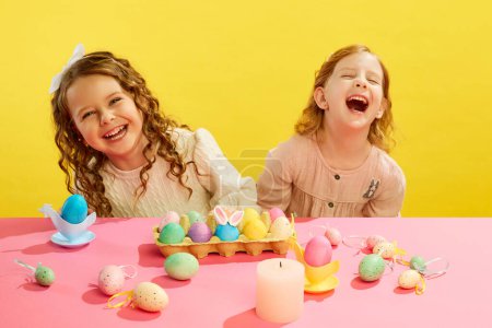 Foto de Niñas, niños riendo alegremente, sentados a la mesa con huevos decorados y pintados sobre fondo amarillo. Concepto de fiesta de Pascua, celebración, tradiciones, infancia, felicidad - Imagen libre de derechos