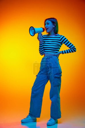 Téléchargez les photos : Portrait complet de la jeune femme habillée pantalon en denim et t-shirt rayé criant bruyamment des informations importantes sur fond dégradé orange. Concept de médias, de mode et de style. Publicité - en image libre de droit