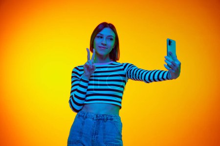 Photo for Half-length portrait of young beautiful girl taking selfie and showing peace gesture to camera against gradient orange background in neon light. Concept of communication, media, information, fashion. - Royalty Free Image
