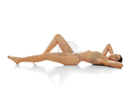 Téléchargez les photos : Belle jeune fille au corps mince couché sur le sol en sous-vêtements sur fond de studio blanc. Épilation, épilation, massage. Concept de beauté naturelle, santé et soins du corps, traitement, cosmétologie - en image libre de droit