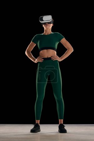 Photo for Full-length image of young woman with slim body in sportswear wearing VR glasses against black studio background. Online sport program. Concept of beauty, health and body care, fitness, innovations - Royalty Free Image