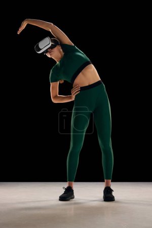 Téléchargez les photos : Jeune femme mince en tenue de sport portant des lunettes VR et de l'entraînement, s'étirant sur fond noir. Entraînement physique virtuel. Concept de sport virtuel, corps et soins de santé, innovations, technologie - en image libre de droit