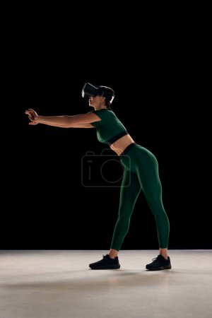 Foto de Servicios innovadores de formación personal. Mujer delgada joven deportiva en entrenamiento de gafas VR, haciendo ejercicios sobre fondo negro. Concepto de deporte virtual, cuerpo, salud, innovaciones, tecnología - Imagen libre de derechos