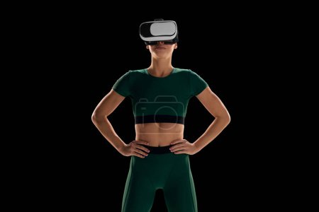 Foto de VR promoción de equipos de fitness y e-sport. Mujer joven delgada que usa gafas VR y ropa deportiva contra fondo de estudio negro. Concepto de deporte virtual, cuidado del cuerpo y la salud, innovaciones, tecnología - Imagen libre de derechos