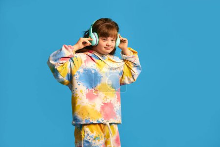 Téléchargez les photos : Adolescente avec le syndrome du duvet dans des vêtements colorés écoutant de la musique dans des écouteurs sur fond bleu studio. Thérapie. Concept d'acceptation, soins, inclusion, santé, diversité, émotions - en image libre de droit