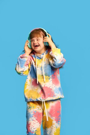 Téléchargez les photos : Joyeux, souriant petite fille avec le syndrome du duvet écouter de la musique dans les écouteurs sur fond bleu studio. Concept d'acceptation, soins, inclusion, santé, diversité, émotions, égalité - en image libre de droit