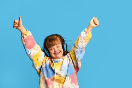 Téléchargez les photos : Joyeux sourire adolescent fille avec le syndrome duvet écouter de la musique dans les écouteurs, exprimant la joie sur fond bleu. Concept d'acceptation, soins, inclusion, santé, diversité, émotions, égalité - en image libre de droit