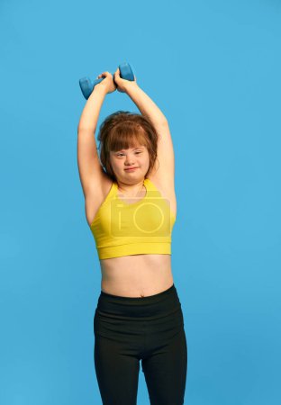 Photo for Teen girl with down syndrome in sportswear training, lifting dumbbells against blue studio background. Concept of acceptance, care, inclusion, health, diversity, emotions, equality - Royalty Free Image