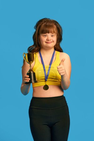Photo for Promotion of equality and acceptance. Teen girl with down syndrome standing with medal and trophy against blue studio background. Concept of sport, care, inclusion, health, diversity, emotions - Royalty Free Image