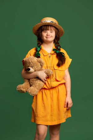Photo for Little girl with down syndrome wearing yellow dress, hat, standing with bear toy against green studio background. Concept of acceptance, care, inclusion, health, diversity, emotions, equality - Royalty Free Image
