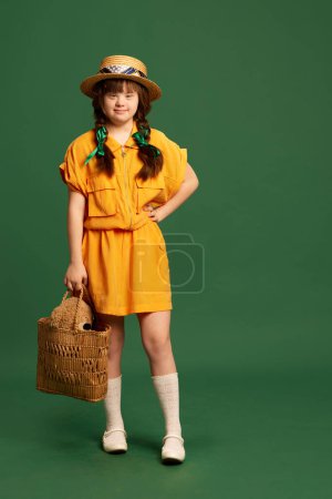 Photo for Beautiful teen girl with down syndrome wearing yellow dress and hat standing against green studio background. Concept of acceptance, care, inclusion, health, diversity, emotions, equality - Royalty Free Image