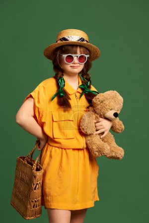 Photo for Portrait of beautiful little girl with down syndrome wearing hat, sunglasses and yellow suit against green background. Concept of acceptance, care, inclusion, health, diversity, emotions, equality - Royalty Free Image