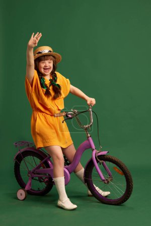 Photo for Happy teen girl with down syndrome wearing yellow suit, sitting on bicycle and having fun over green studio background. Concept of acceptance, care, inclusion, health, diversity, emotions, equality - Royalty Free Image