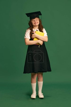 Photo for Inclusive education campaign. Girl with down syndrome standing with books, wearing graduation cap against green studio background. Concept of acceptance, care, inclusion, health, education, equality - Royalty Free Image